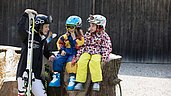 Kinder sitzen mit Ski-Ausrüstung auf einem Holzstamm und freuen sich auf die Piste im Familienhotel Bavaria im Allgäu.