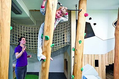 Kind beim Klettern an einer Holzwand im Indoor-Spielbereich des Familienhotels Amiamo im Salzburger Land, beaufsichtigt von einer lächelnden Betreuerin, die ein aktives und kinderfreundliches Hotelkonzept hervorhebt.
