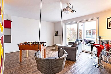 Geräumiger Freizeitraum im Familienhotel Deichkrone mit einem Tischfußball, bequemen Sitzgelegenheiten, einem Projektor und Zugang zu einer Terrasse, ideal für Teenager und Familienunterhaltung.