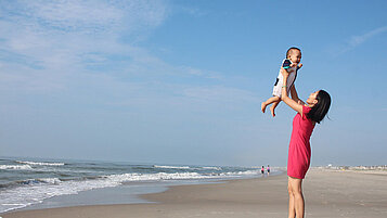 Mutter steht mit ihrem Baby am Strand und hebt es in die Höhe.