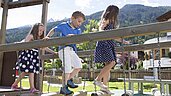 Drei Kinder spielen im Sommer auf dem Outdoor-Spielplatz des Familienhotels Alpenhotel Kindl in Tirol. Auf dem Outdoor Spielplatz hat man einen herrlichen Blick auf die umliegenden Berge.