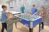 Kinder verschiedener Altersgruppen haben Spaß beim Spielen an einem farbenfrohen Tischfußball in einem Spielzimmer, dessen Wände mit Worten wie 'joy' und 'happy' dekoriert sind, was eine fröhliche und energiegeladene Atmosphäre schafft.