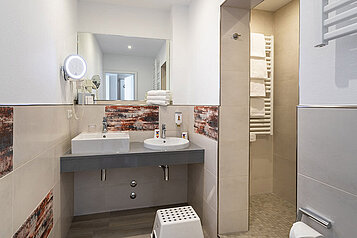 Modernes Badezimmer im Familienhotel Deichkrone an der Nordsee, ausgestattet mit einem Doppelwaschtisch, großem Spiegel, Handtuchheizkörper und dekorativen Fliesen mit Rosteffekt für ein warmes Ambiente.