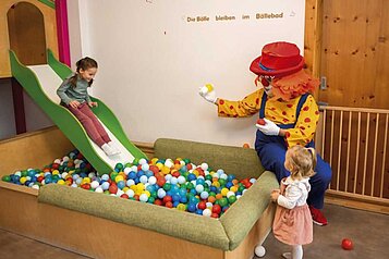 Kind rutscht auf einer Kinderrutsche ins Bällebad des Happy-Clubs im Familienhotel Alpenhotel Kind in Tirol. Auch Clown Happy, das Maskottchen des Familienhotels, spielt mit.
