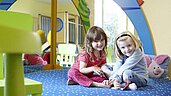 Zwei lächelnde Kinder spielen in einem farbenfrohen Indoor-Spielbereich, umgeben von weichen Polstern und Spielsachen, was die kinderfreundlichen Einrichtungen des Familienhotels Deichkrone an der Nordsee hervorhebt.