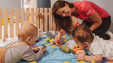Zwei Babys liegen gemeinsam mit einer Babybetreuerin des Familienhotels Huber in Südtirol auf einer Spieldecke und beschäftigen sich mit altersgerechten Spielsachen.