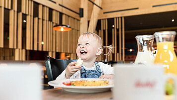 Ein Baby sitz am Tisch beim Essen und lacht und hat ein Stück Brot in der Hand.