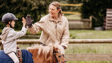 Kind beim Reiten eines Ponys gibt einer Betreuerin im Landhaus Averbeck fröhlich ein High-Five.