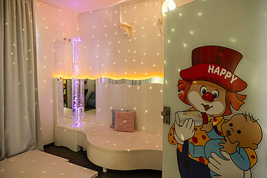 Entspannendes Snoezelen-Zimmer für Kinder im Familienhotel Schreinerhof mit weichen Sitzgelegenheiten, Sternenhimmelbeleuchtung und einer leuchtenden Wassersäule, neben einer Tür mit einem freundlichen Clown-Motiv.