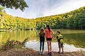 Drei Kinder stehen vor einem See und Blicken auf den See und den umliegenden Wald im Sommer.