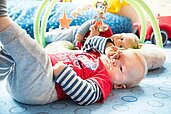 Babybereich im Familienhotel Deichkrone mit einem lächelnden Baby, das auf einer bunten Spielmatte mit Spielbogen und weichen Spielzeugen liegt, ein einladender und sicherer Ort für die kleinsten Gäste.