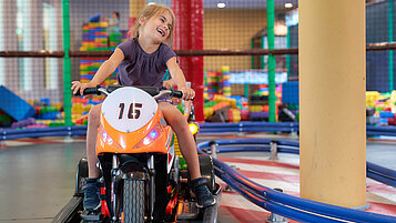 Ein fröhliches Mädchen fährt ein elektrisches Kinder-Motorrad auf einer Indoor-Rennbahn im Familienhotel Schreinerhof im Bayrischen Wald, mit einem bunten Spielbereich im Hintergrund.