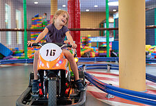 Ein fröhliches Mädchen fährt ein elektrisches Kinder-Motorrad auf einer Indoor-Rennbahn im Familienhotel Schreinerhof im Bayrischen Wald, mit einem bunten Spielbereich im Hintergrund.