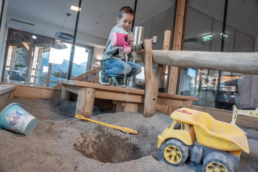 Ein Kind spielt mit einer Gießkanne im Indoor-Matschplatz im Almfamilyhotel Scherer in Tirol.