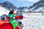 Familie macht eine Pause vom Skifahren und sitzt im Schnee bei Sonnenschein.