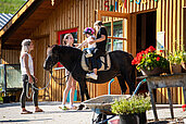 Zwei Kinder sitzen vor der Reithalle des Familienhotels Der Böhmerwald auf einem Pferd. Die Mutter führt das Pferd mit den beiden Kindern.