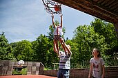 Vater hält seine Tochter in die Luft, damit diese einen Basketball auf dem Basketballplatz des Kinderhotels Bruckwirt in den Korb werfen kann.