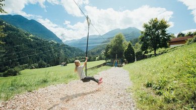Mädchen sitzt auf einer Schaukel auf dem Outdoor-Spielplatz des Familienhotels Sailer. Der Spielplatz ist umgeben von Natur.
