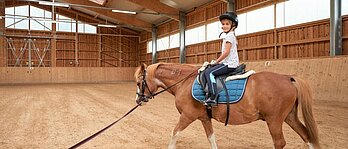 Kind sitzt beim Reitunterricht in der Reithalle des Familienhotels Schreinerhof auf dem Pferd.