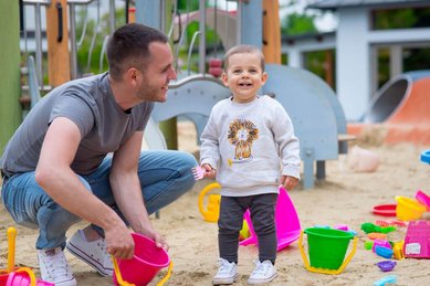 Spielspaß im Sand: Vater und Kleinkind genießen gemeinsam den Spielplatz im Kolping Family Resort in Ungarn, dem perfekten Familienhotel für unvergessliche Urlaubsmomente.