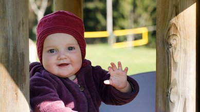 Ein Baby mit lila Jacke und Mütze winkt fröhlich in die Kamera auf dem Außengelände des Familienhotels Ramsi in Kärnten