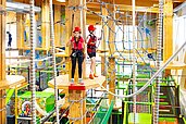 Zwei Kinder in Sicherheitsausrüstung gehen über Holzplattformen in einem bunten Indoor-Hochseilgarten im Hotel Feldberger Hof, umgeben von Seilen und Netzen.