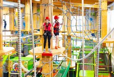 Zwei Kinder in Sicherheitsausrüstung gehen über Holzplattformen in einem bunten Indoor-Hochseilgarten im Hotel Feldberger Hof, umgeben von Seilen und Netzen.
