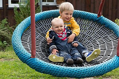 Zwei Kinder sitzen in einer Nestschaukel im Aussenbereich des Familienhotels Alphotel