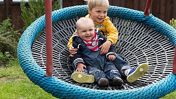 Zwei Kinder sitzen in einer Nestschaukel im Aussenbereich des Familienhotels Alphotel