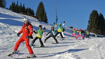 Kind mit Helm und Skiausrüstung steht auf verschneiter Piste vor einer malerischen Bergkulisse, was Wintersportaktivitäten für Kinder in einem familienfreundlichen Hotelumfeld veranschaulicht.