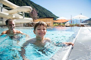 Junge spielt mit Vater am Beckenrand des Outdoor-Schwimmbeckens im Familienhotel Huber in Südtirol.