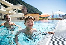 Junge spielt mit Vater am Beckenrand des Outdoor-Schwimmbeckens im Familienhotel Huber in Südtirol.