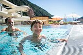 Junge spielt mit Vater am Beckenrand des Outdoor-Schwimmbeckens im Familienhotel Huber in Südtirol.