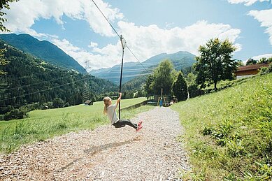 Mädchen sitzt auf einer Schaukel auf dem Outdoor-Spielplatz des Familienhotels Sailer. Der Spielplatz ist umgeben von Natur.