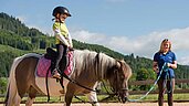 Kleines Mädchen sitzt beim Reitunterricht im Spa- & Familien-Resort Krone auf dem Pferd und wird an der Lounge von einer Reitlehrerin geführt.