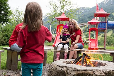 Eine Mutter sitzt mit ihren Kindern am Lagerfeuer und macht Stockbrot mit den Kindern im Familienhotel Bavaria im Allgäu