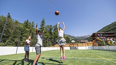 Kinder spielen auf dem Spielplatz des Familienhotels Galtenberg Family & Wellness Resort Basketball bei gutem Sommer Wetter.