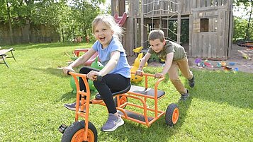 Lachendes Mädchen sitzt auf einem Dreirad und wird von einem Jungen angeschoben. Im Hintergrund ist der Außenspielplatz des Familienhotels Rhön Feeling zu sehen.