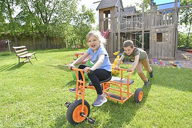 Lachendes Mädchen sitzt auf einem Dreirad und wird von einem Jungen angeschoben. Im Hintergrund ist der Außenspielplatz des Familienhotels Rhön Feeling zu sehen.