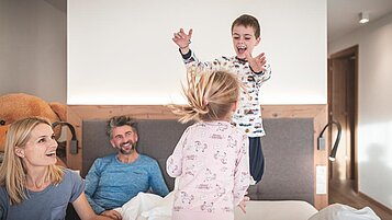 Vierköpfige Familie tobt auf dem Bett im Familienzimmer des Familienhotels Alpenhof Dolomit Family in Südtirol.