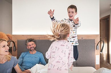 Vierköpfige Familie tobt auf dem Bett im Familienzimmer des Familienhotels Alpenhof Dolomit Family in Südtirol.