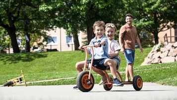 Zwei Kinder fahren gemeinsam ein Dreirad, während ein Erwachsener im Familotel-Garten hinterherläuft.