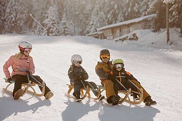 Eine Familie hat Spaß beim Rodeln auf einer sonnenbeschienenen Piste im Winter am Hotel Habachklause im Salzburger Land, umgeben von malerischen Schneelandschaften.