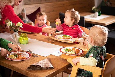 Mutter sitzt mit vier Kleinkindern am Tisch im familienfreundlichen Restaurant des Familienhotels Landgut Furtherwirt in Tirol. Die Kinder sitzen im Hochstuhl und essen Fischstäbchen mit Kartoffeln vom bunten Kinderteller.