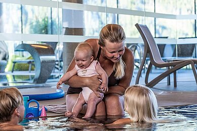 Eine Mutter sitz am beckenrand des Pools und hält ihr Baby im arm und unterhält sich mit zwei anderen Kindern im Pool des Familienhotels Scherer.