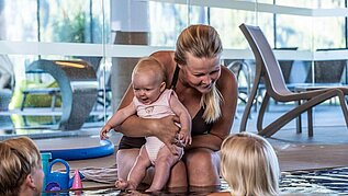 Eine Mutter sitz am beckenrand des Pools und hält ihr Baby im arm und unterhält sich mit zwei anderen Kindern im Pool des Familienhotels Scherer.