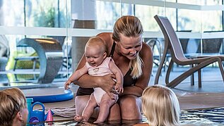 Eine Mutter sitz am beckenrand des Pools und hält ihr Baby im arm und unterhält sich mit zwei anderen Kindern im Pool des Familienhotels Scherer.