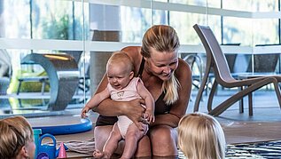 Eine Mutter sitz am beckenrand des Pools und hält ihr Baby im arm und unterhält sich mit zwei anderen Kindern im Pool des Familienhotels Scherer.