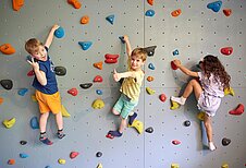 Drei Kinder klettern die Kletterwand in der Indoor-Spielhalle vom Familienhotel Das Bayrischzell in Oberbayern hoch.