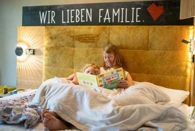 Mutter und Tochter kuscheln gemeinsam im gemütlichen Bett auf ihrem Familienzimmer im Familienhotel im Bayerischen Wald und lesen ein Buch